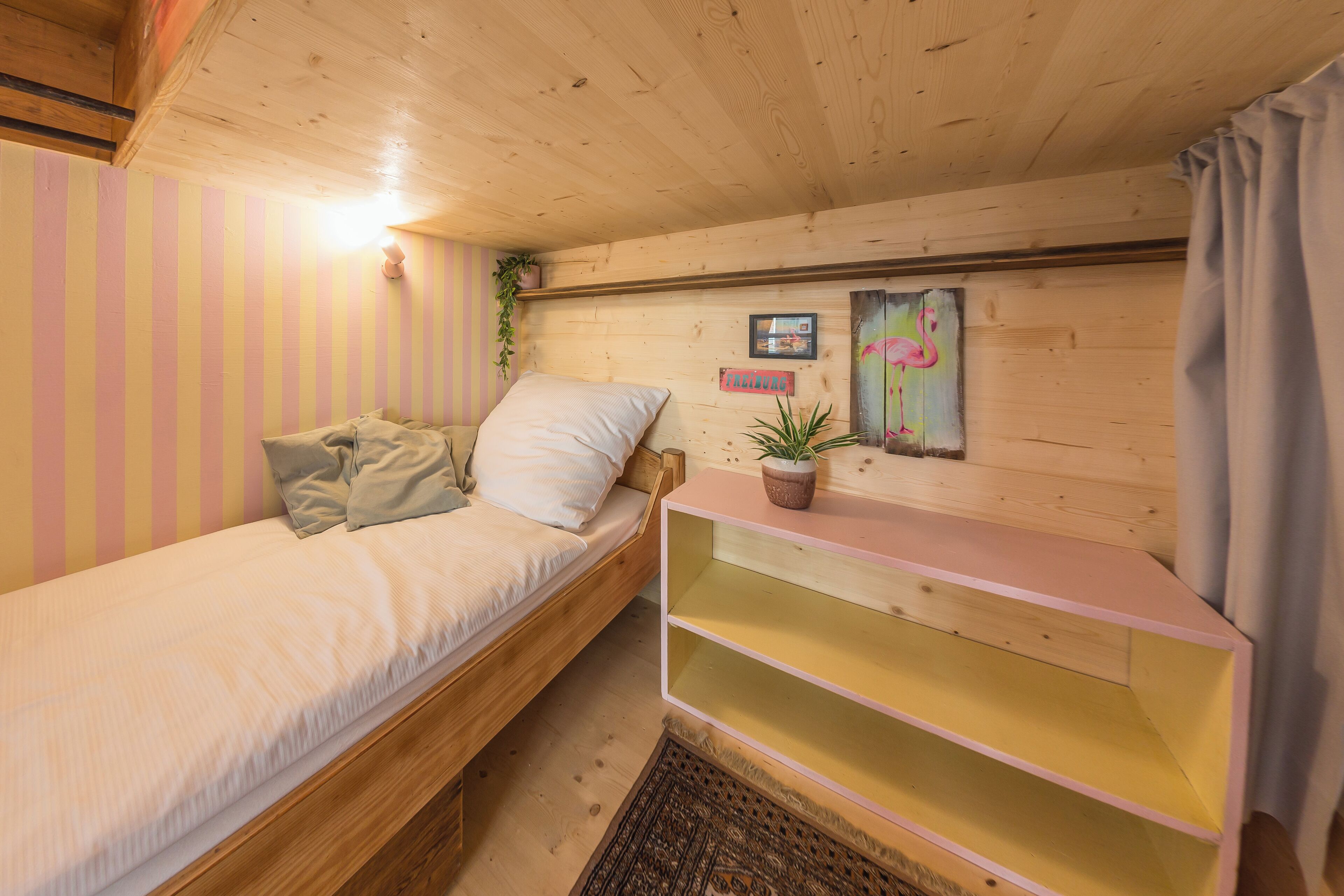 Ein gemütliches Zimmer mit einem Holzbett, einer kleinen Ablagefläche mit einer Pflanze und einem an der Wand hängenden Bild.