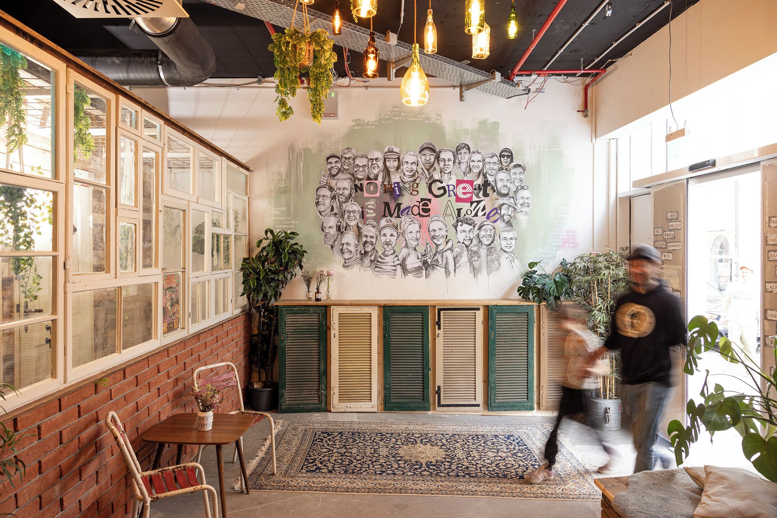 A cafe with a brick wall, wooden tables, and chairs, a mural with faces, and hanging plants.