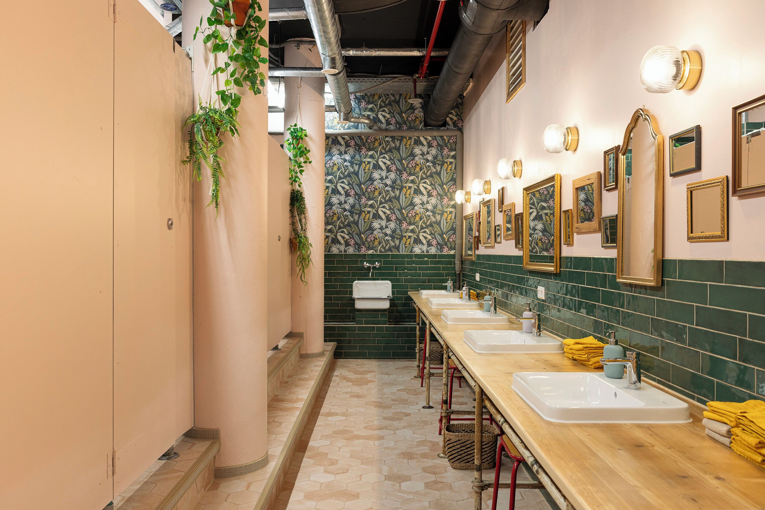 A long, narrow bathroom with multiple sinks, green tiled walls, and hanging plants.