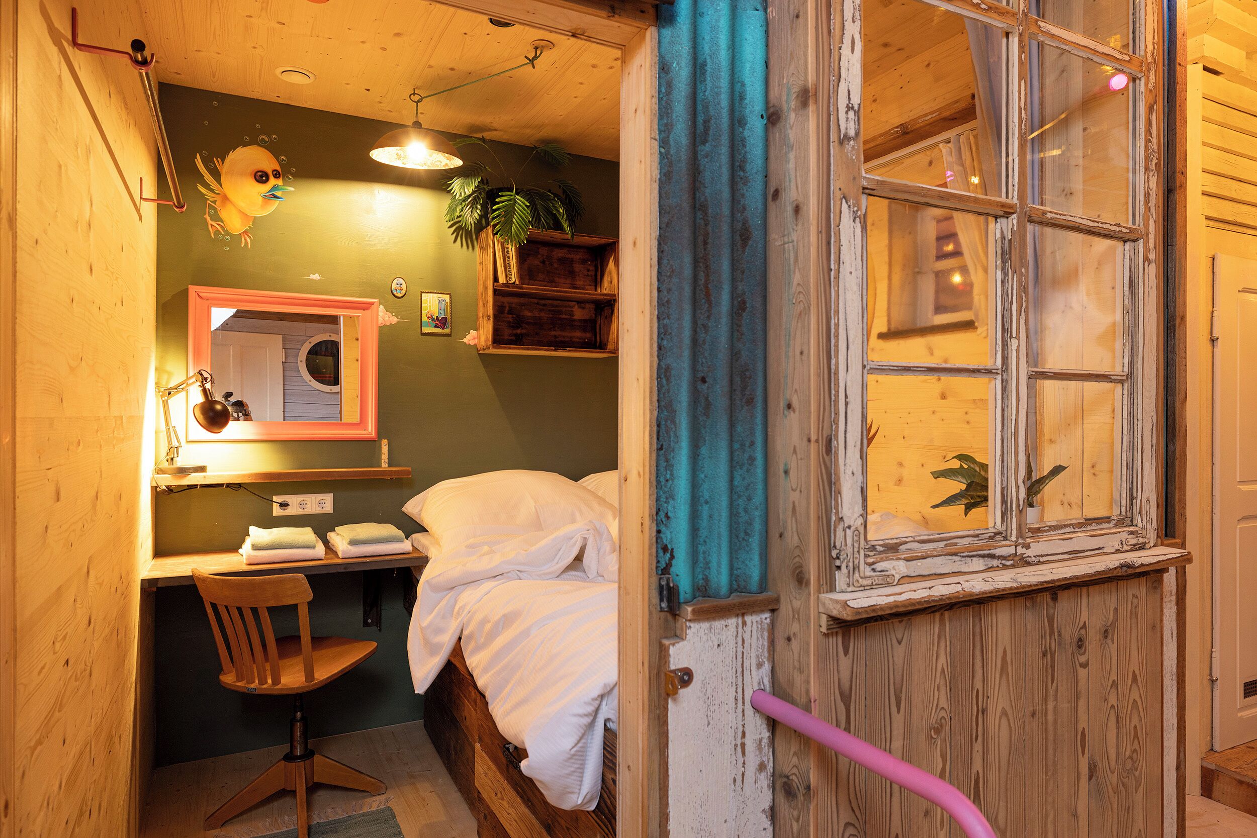 A cozy bedroom with a wooden bed, a desk with a chair, a window with a curtain, and a wall-mounted shelf with plants.