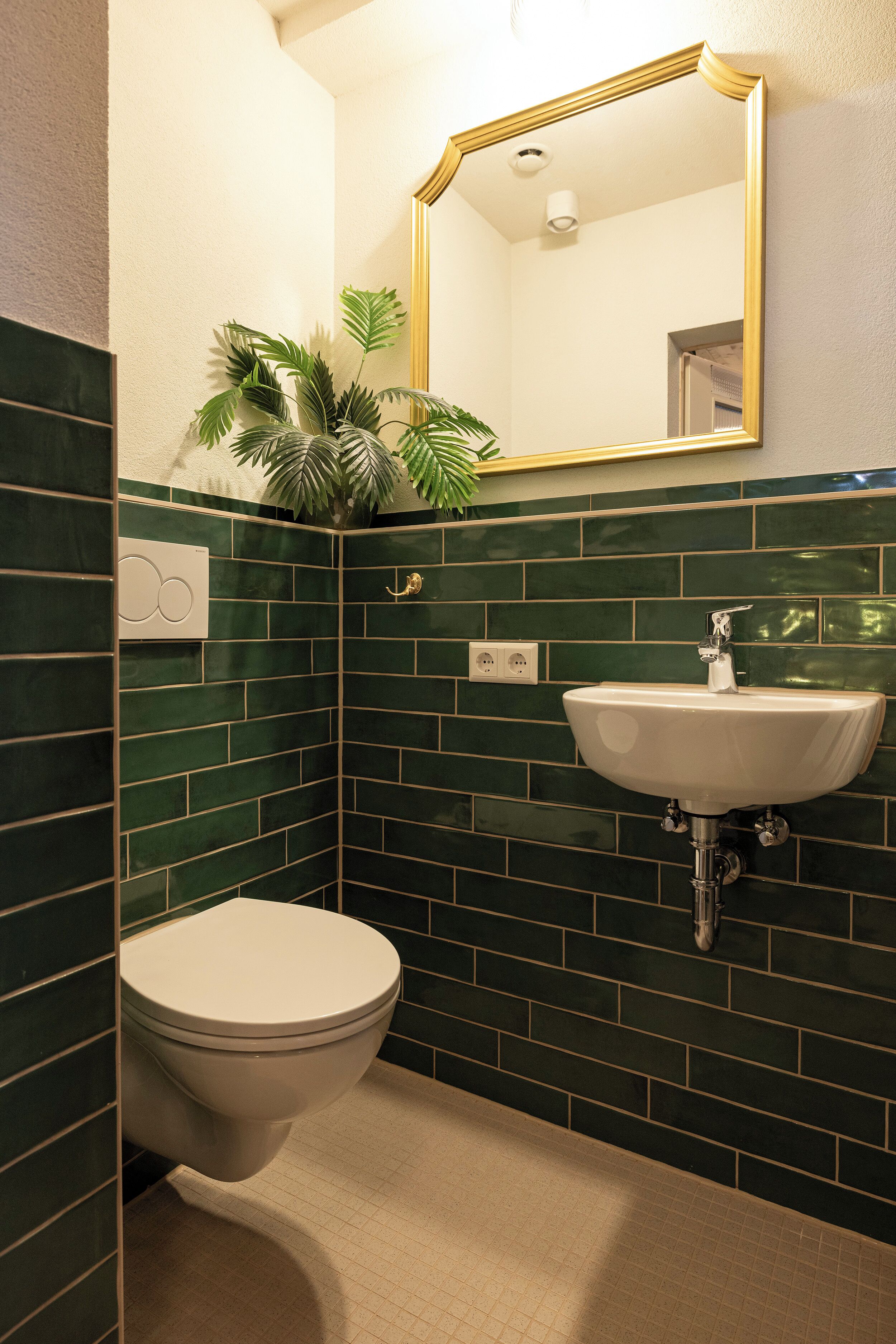 Une salle de bain avec des toilettes blanches, des murs recouverts de carreaux verts, un lavabo blanc et un miroir avec un cadre doré.