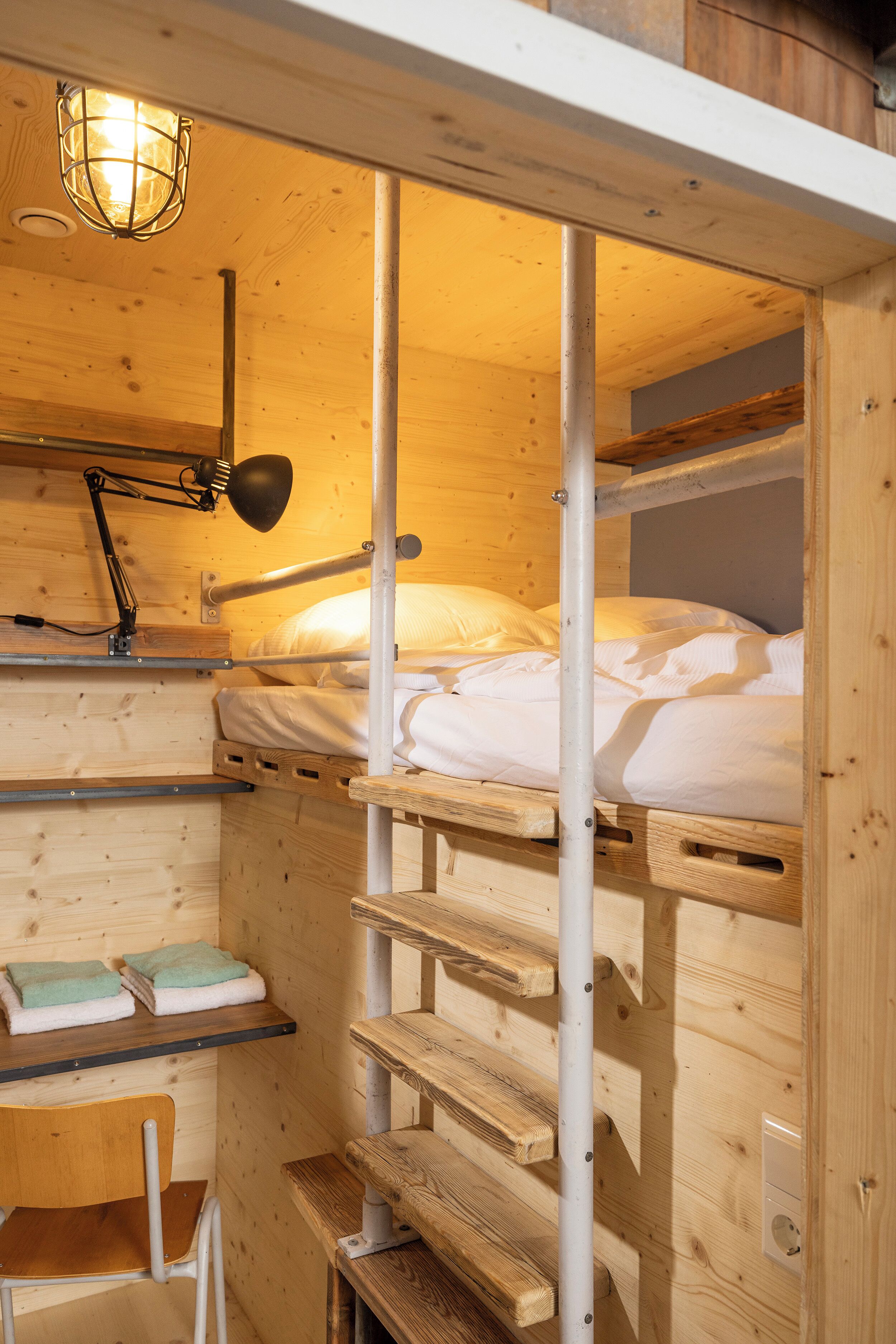 A bunk bed room with a desk and chair, a lamp, and a shelf with towels.