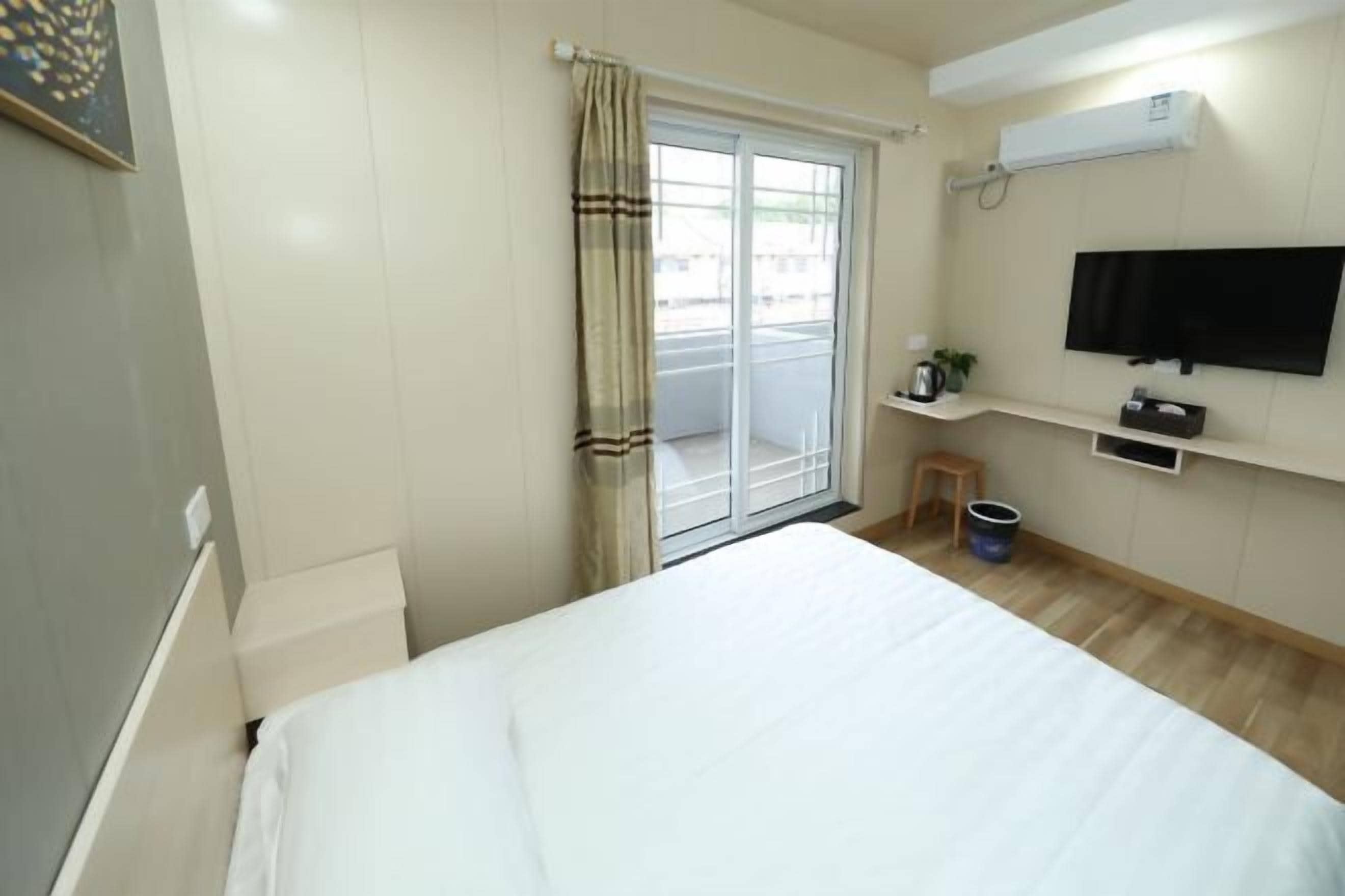 Quarto de hotel com uma cama, televisão na parede, uma pequena mesa com uma planta e uma janela com cortinas.