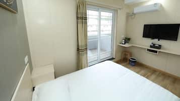 Quarto de hotel com uma cama, televisão na parede, uma pequena mesa com uma planta e uma janela com cortinas.