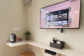 A hotel room with a flat-screen TV mounted on the wall displaying a menu, a small table with a kettle and cups, and a plant on the wall. - Kunxin Hostel (Shanghai)