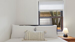 Una cama bien hecha con almohadas blancas y a rayas, una lámpara de noche y una ventana con vista a árboles y un muro de ladrillo.