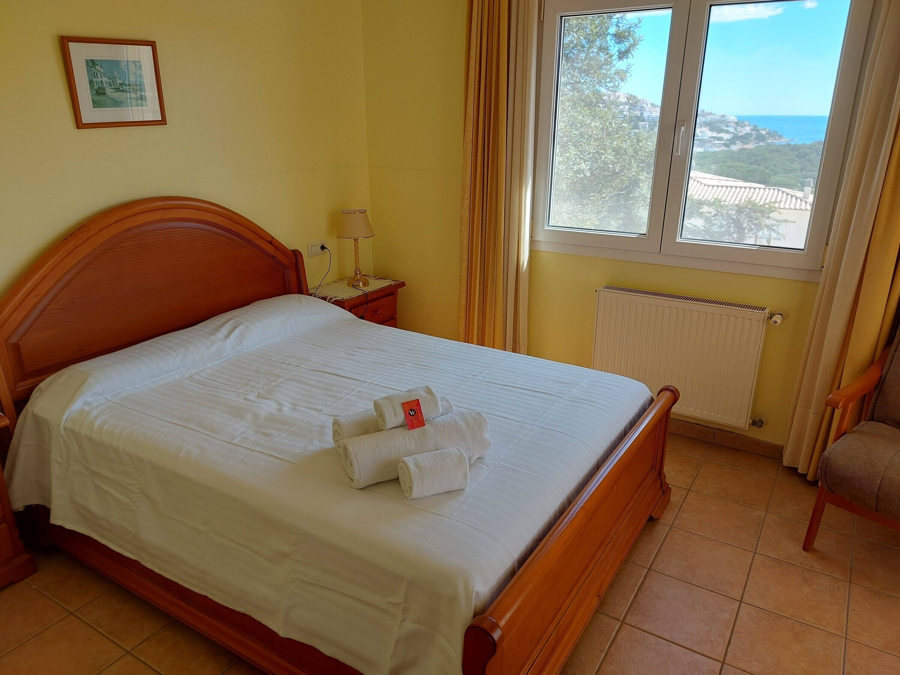 Una cama bien hecha con una colcha blanca y dos toallas enrolladas, un cabecero de madera, una mesita de noche con una lámpara, un cuadro en la pared y una ventana con vistas al paisaje costero.