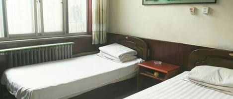 A room with two single beds, a wooden headboard, a small bedside table, and a window with curtains.