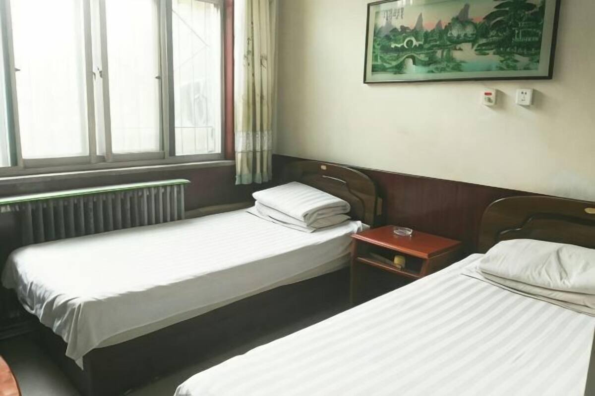A hotel room with two single beds, a wooden headboard, a small bedside table with a lamp, a window with curtains, and a painting on the wall.