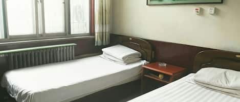 A hotel room with two single beds, a wooden headboard, a small bedside table with a lamp, a window with curtains, and a painting on the wall.