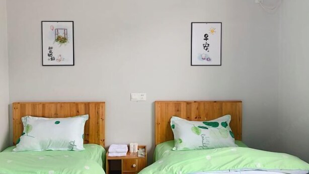 A hotel room with two beds, each with a wooden headboard and green bedding. There are two framed pictures on the wall, one with a plant and the other with a sun. - Xiaotaoyuan Inn (Wuyuan)