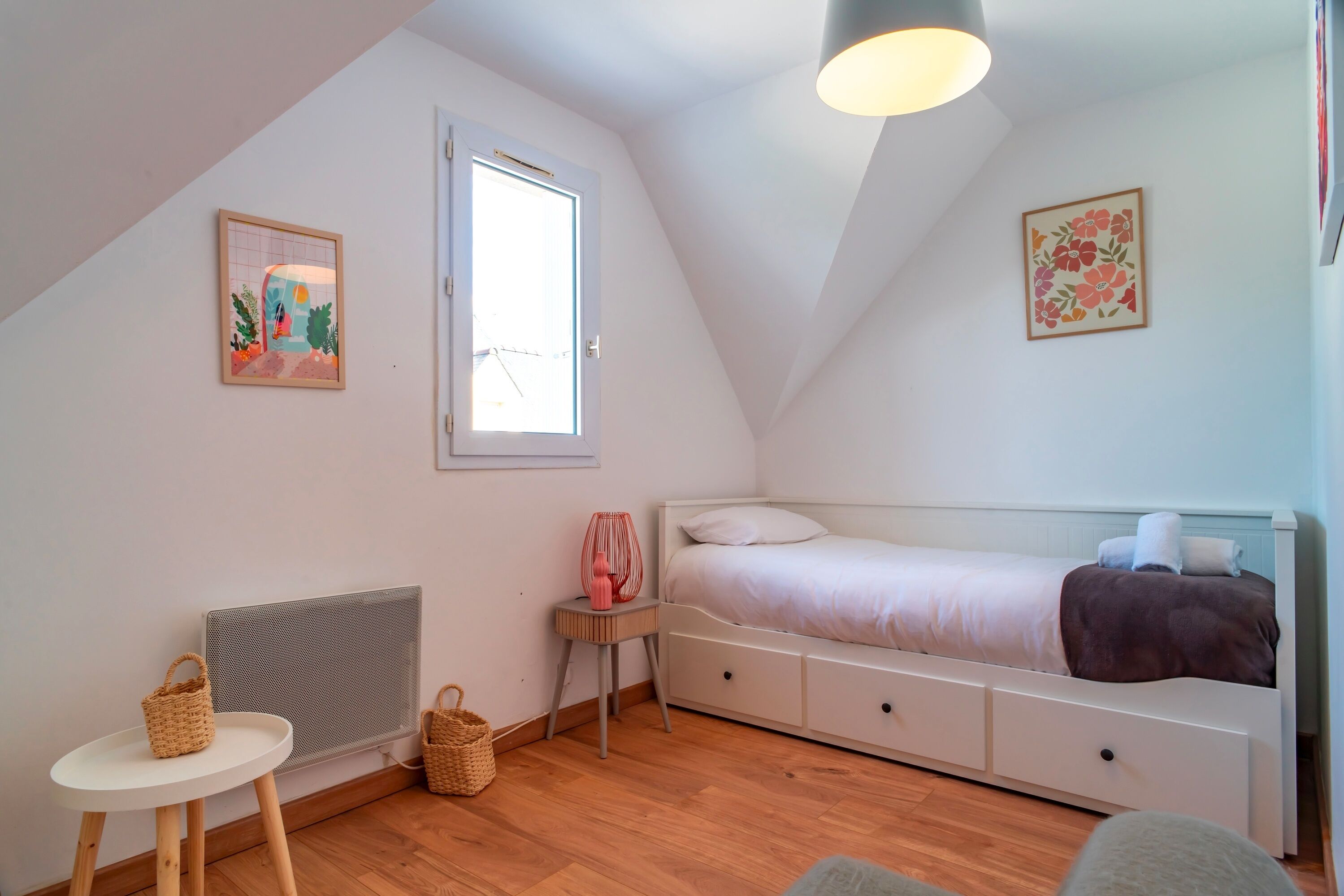 Une chambre avec un plafond en pente, un lit, un radiateur, une petite table et deux paniers fixés au mur.
