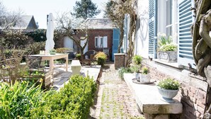 Un jardín con un sendero de ladrillo que lleva a una mesa y sillas de madera, rodeado de plantas en macetas y un edificio azul con contraventanas.