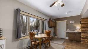A dining area with a round table and chairs, a window with curtains, a ceiling fan, and a kitchen area with wooden cabinets and a welcome sign. - Emily Channel Cabin on the water with ATV trail access from your doorstep! (Emily)