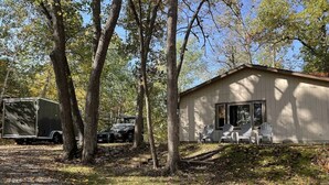 A cabin with a covered porch, a trailer, and a vehicle parked in a wooded area. - Emily Channel Cabin on the water with ATV trail access from your doorstep! (Emily)