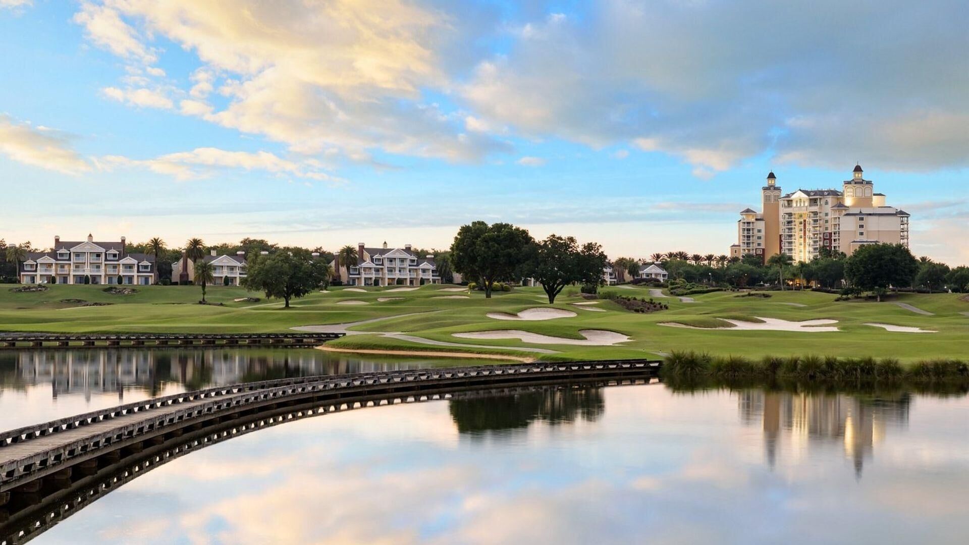A golf course with a water body in the foreground, a bridge, and a large hotel with multiple buildings in the background.