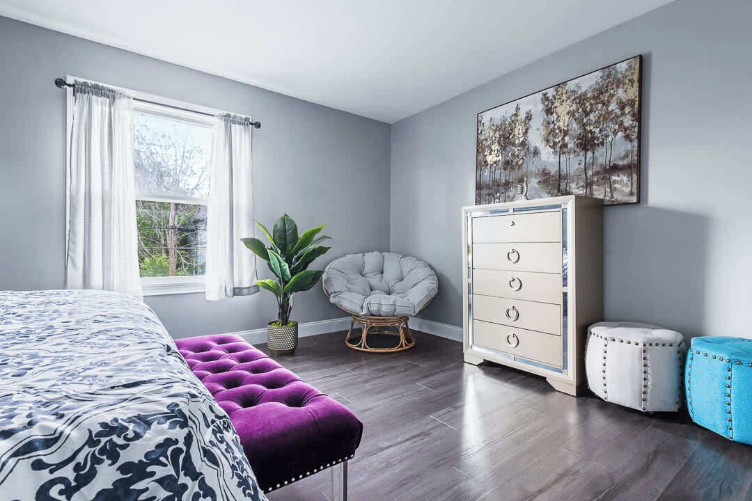 A bedroom with a bed, a dresser, a chair, a plant, and a painting on the wall.
