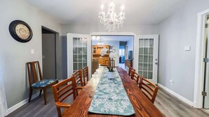 Een eetruimte met een lange houten tafel, stoelen, een kroonluchter en een klok aan de muur.