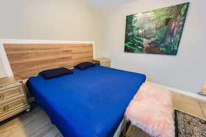 Een slaapkamer met een houten hoofdbord, een blauwe spreideken, een roze zachte plaid, een nachtkastje, een lade en wanddecoratie met een bosmotief.