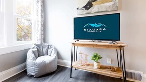 Huoneessa on harmaa papupussi-istuin, puinen TV-taso, jossa on Niagara Hospitality -logo, ikkuna, jossa on verhot, ja seinälle ripustettu abstrakti maalaus.