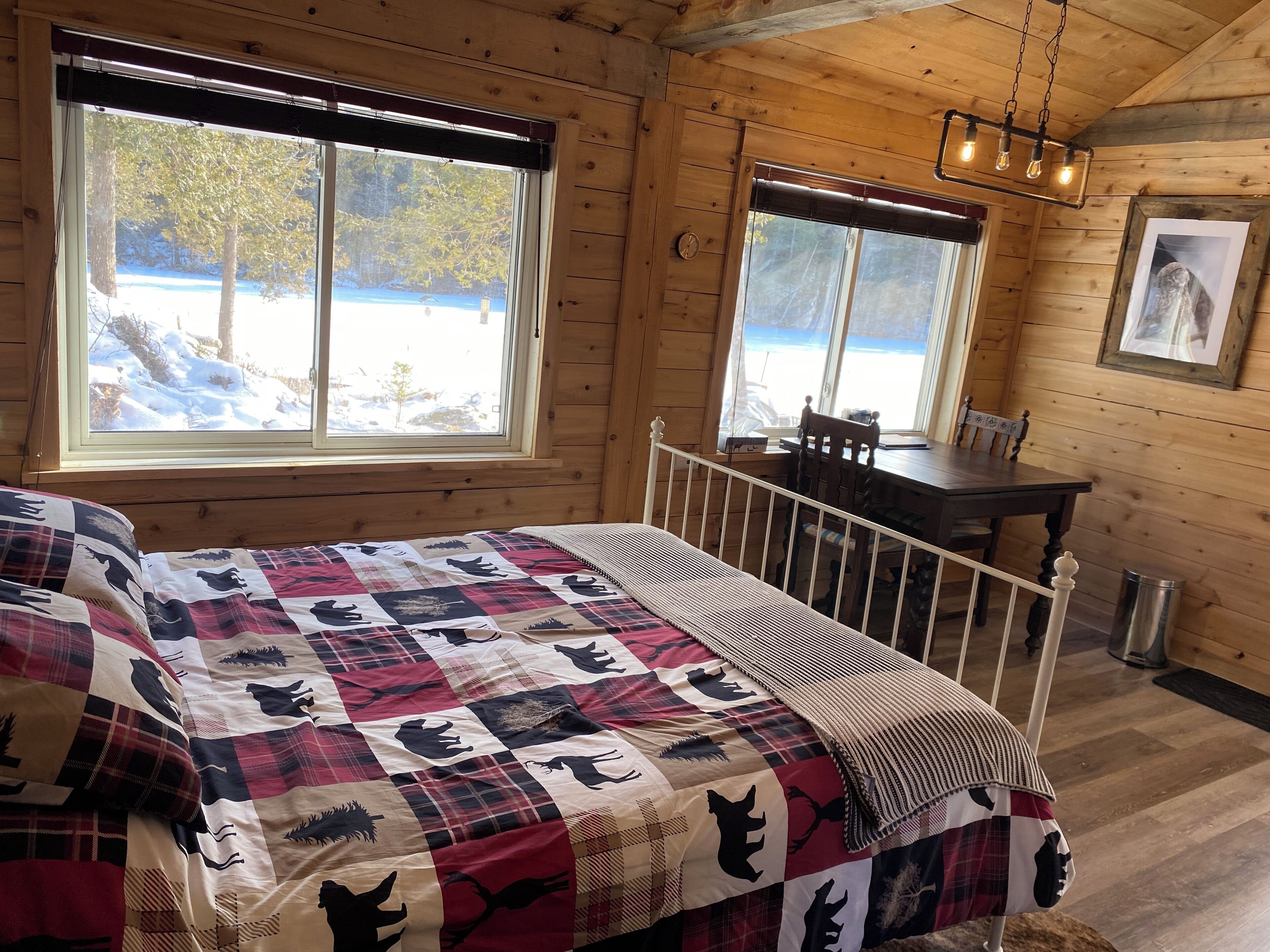 Ein gemütliches Blockhäuschen-Zimmer mit einem großen Bett, einem hölzernen Schreibtisch und Blick auf eine verschneite Landschaft durch zwei Fenster.