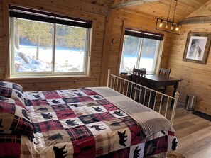 Une chambre confortable dans un chalet, avec un grand lit, un bureau en bois et une vue sur un paysage enneigé par deux fenêtres.