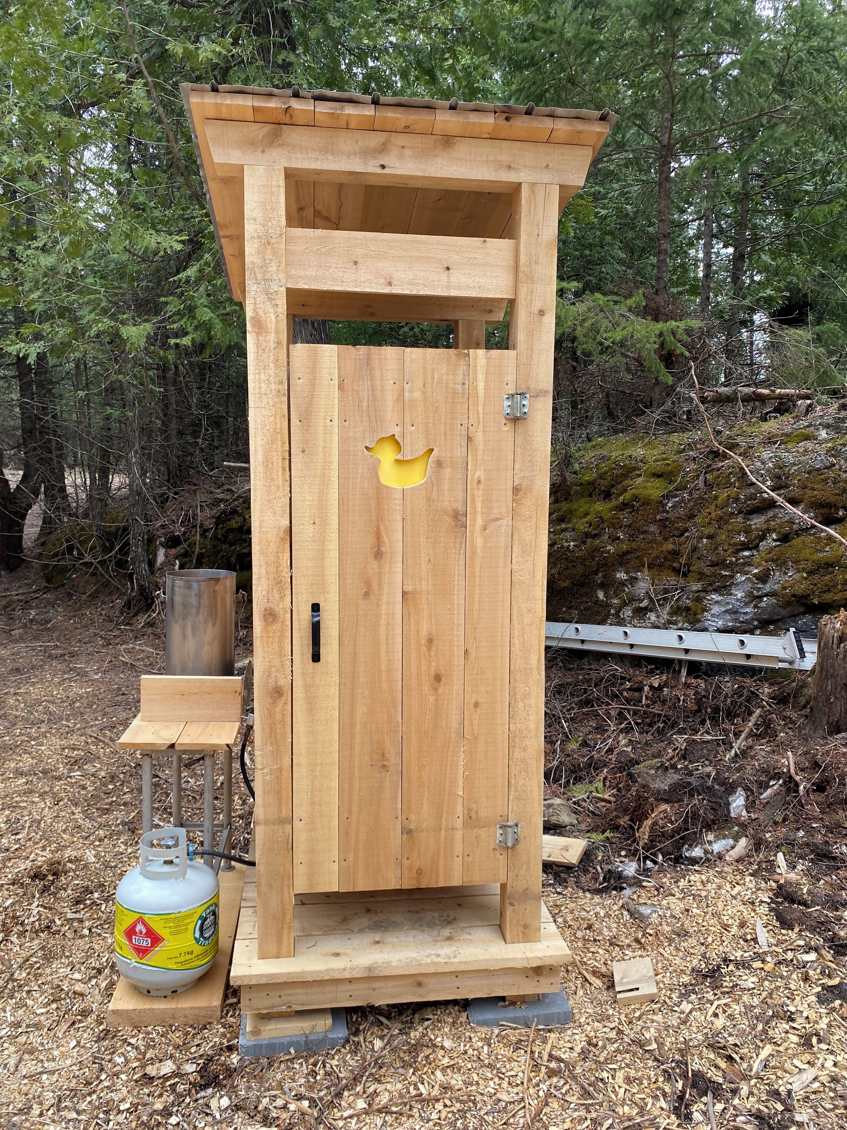 En liten utedass av trä med en gul målad anka som dekoration, en metalldörr och en gastank i närheten.