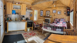 A cabin interior with a kitchen, a bed with a plaid bedspread, a rocking chair, and a fireplace.