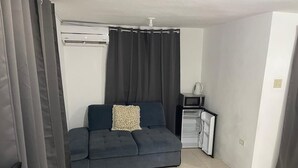 A compact living space with a dark-colored sofa, a patterned pillow, a small table with a kettle, and a white air conditioning unit. - 6-bedroom studio in welcoming Ocho Rios with WiFi, AC Escape to Tranquility (Ocho Rios)