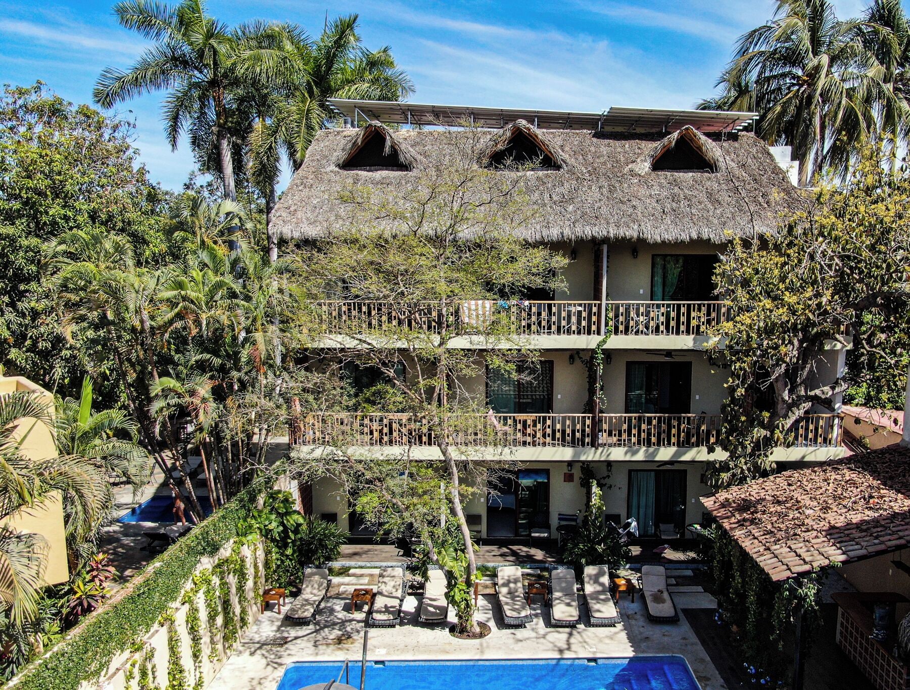 Edificio de dos pisos con piscina, rodeado de abundante vegetación y palmeras.