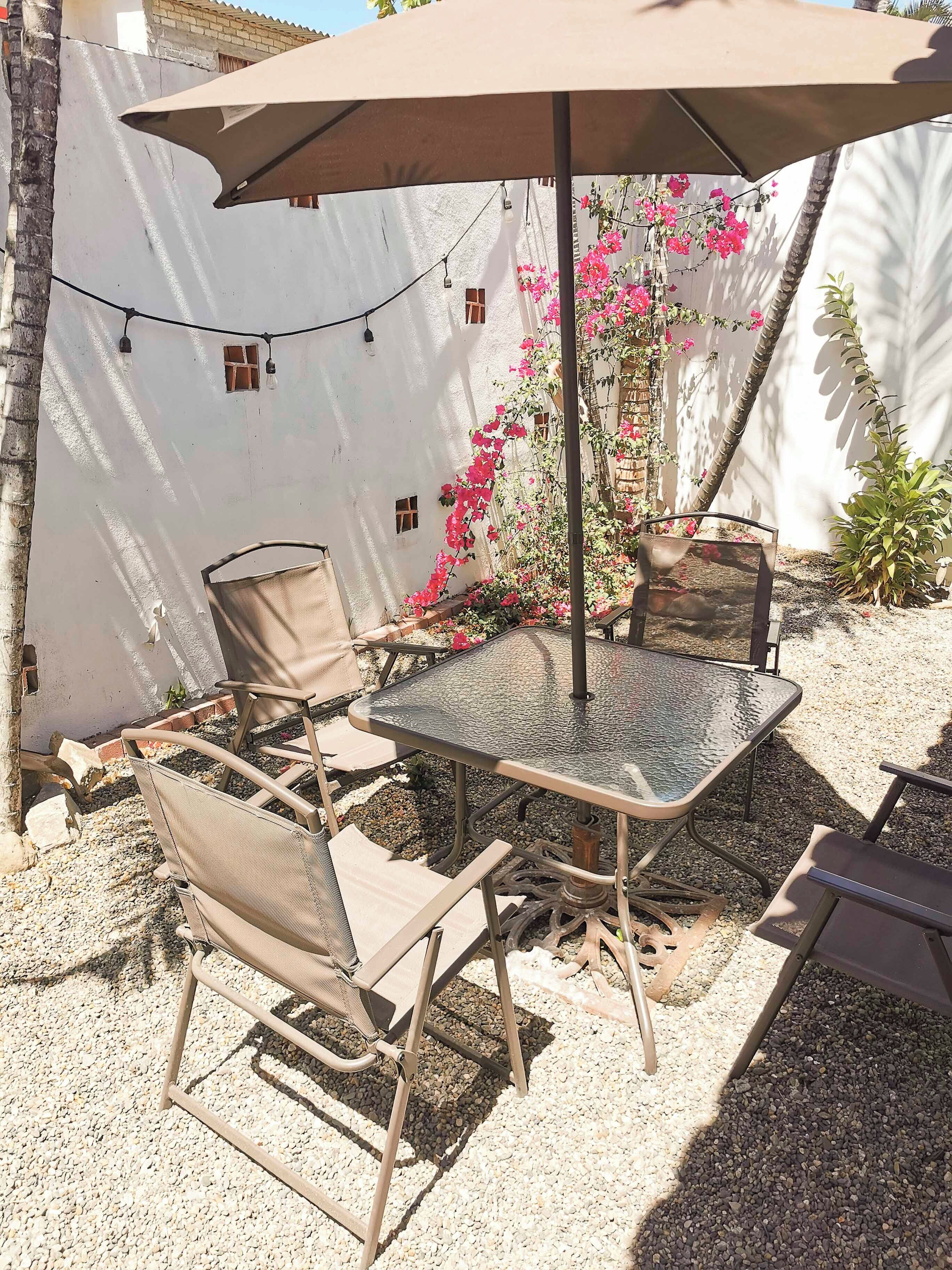 Un patio al aire libre con una mesa de vidrio, sillas de metal, un paraguas grande y luces decorativas tipo guirnalda.