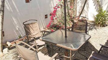 Un patio al aire libre con una mesa de vidrio, sillas de metal, un paraguas grande y luces decorativas tipo guirnalda.