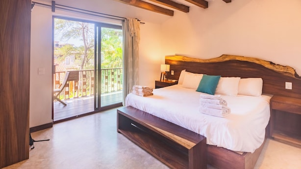 Quarto moderno com uma cama grande, cabeceira de madeira, mesa de cabeceira, luminária e vista para uma varanda com grade de madeira.
