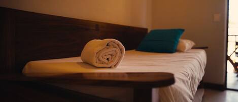 A neatly made bed with a rolled towel, a teal pillow, and a wooden headboard.