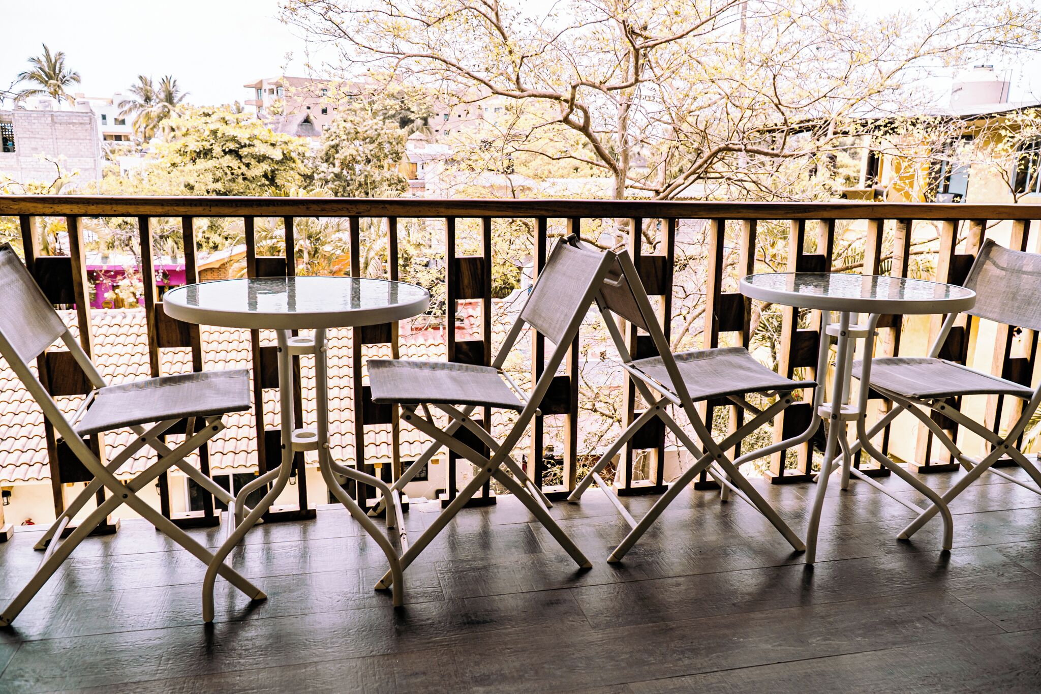 A balcony with four tables and chairs, overlooking a residential area with trees and buildings.