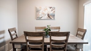 A dining area with a wooden table and chairs, a painting on the wall, and a potted plant on the table.