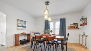 Un comedor con una mesa de madera, seis sillas, un tapete, un escritorio con una computadora, una planta y un colgador de luz.