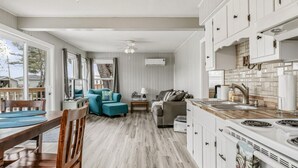 Cozinha moderna com armários brancos, bancada de madeira e fogão embutido. Há uma área de jantar com mesa e cadeiras de madeira, e uma sala de estar com sofá e mesa de centro.