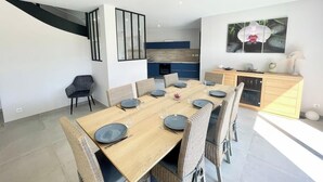 A modern dining area with a wooden table set for six, surrounded by wicker chairs. A kitchen with blue cabinets is visible in the background. - Luxury Maison d'Architecte neuve de standing (Larmor-Baden)