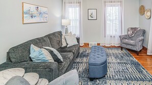 Una sala de estar con un sofá gris, un reposapiés azul, un sillón gris y un tapete estampado. Hay dos ventanas con cortinas translúcidas, una lámpara de pie y dos obras de arte colgadas en la pared.