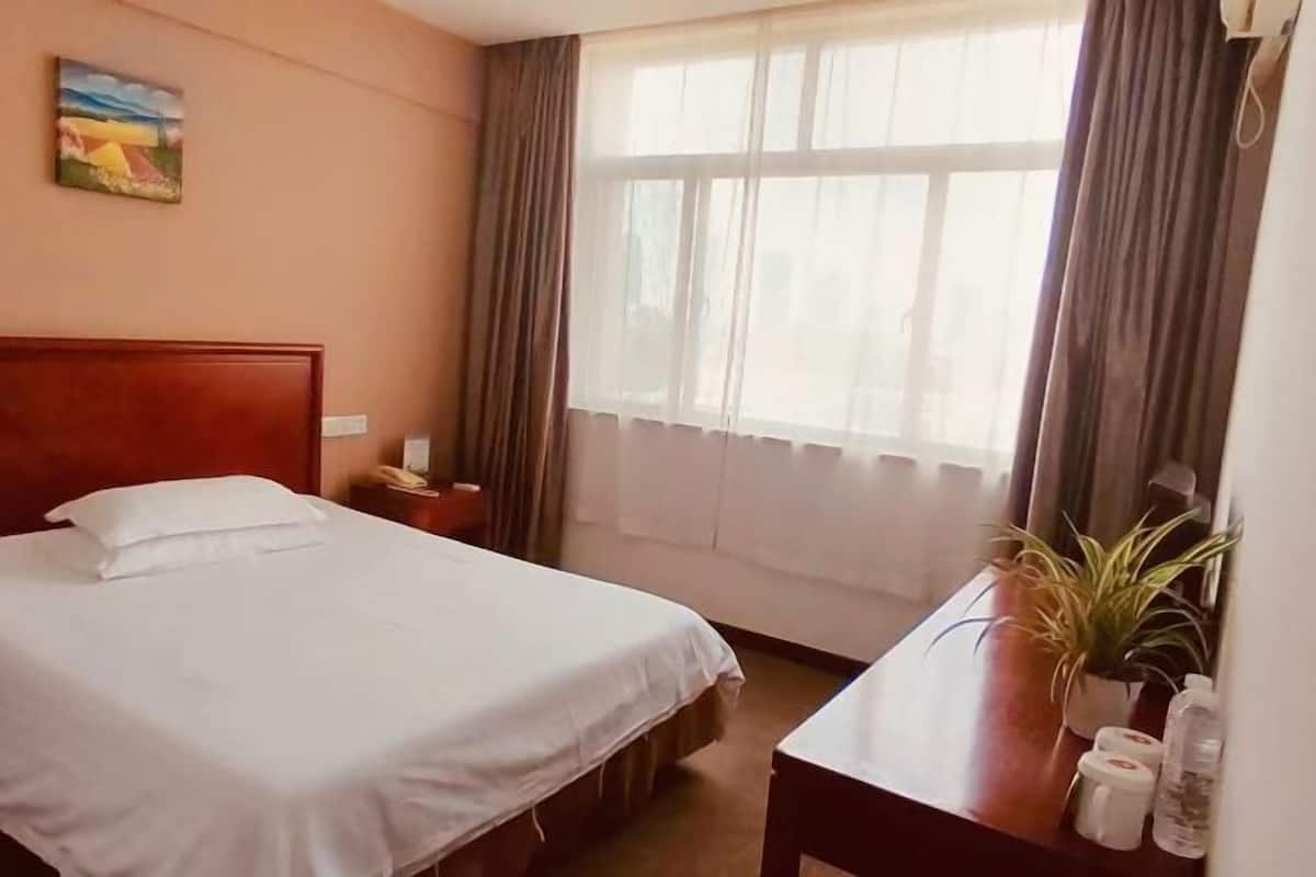 A hotel room with a single bed, a bedside table, a nightstand, a window with curtains, and a painting on the wall.