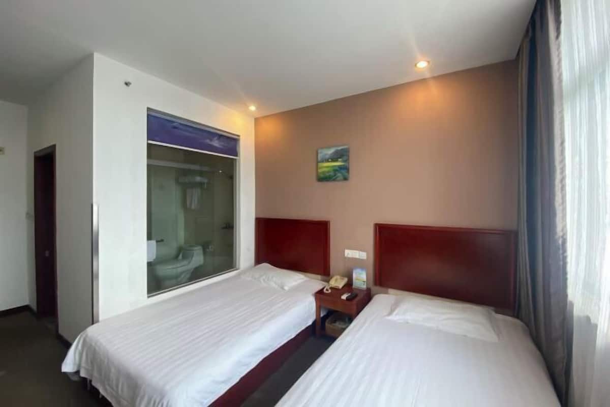 A hotel room with two single beds, a large window, a painting on the wall, and a bedside table with a phone.