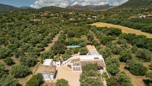 An aerial view of a property with a central building surrounded by dense vegetation, a swimming pool, and a hilly landscape in the background.