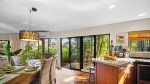 A modern kitchen with a dining area, large windows, and a view of greenery.