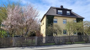 Une maison à deux étages, avec un toit sombre et des murs clairs, entourée d’une clôture en bois et d’arbres en fleurs.