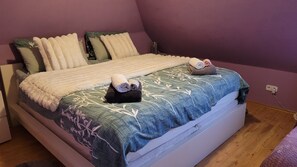 En seng med et mønstret sengetæppe, puder og to sammenrullet håndklæder.