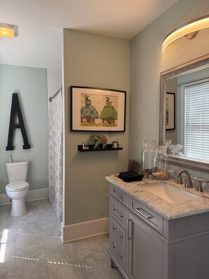 A modern bathroom with a toilet, sink, and shower area. There is a framed picture on the wall and a shelf with decorative items.
