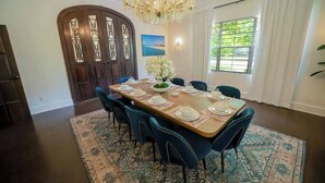 Une salle à manger avec une grande table en bois, dressée pour six personnes, entourée de chaises recouvertes d’un tissu bleu. Un lustre est suspendu au plafond, et une fenêtre avec des rideaux blancs est visible.
