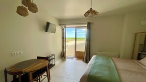 Een hotelkamer met een bed, een bureau en een televisie. Er is een schuifdeur naar een balkon met uitzicht op het strand.