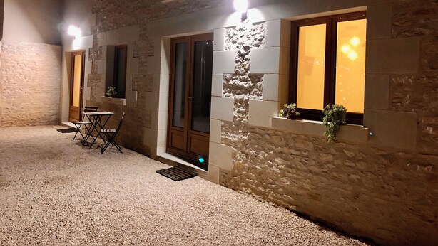 Een gebouw met stenen muren, een houten deur en ramen met verlichting. Buiten staan een kleintje tafeltje en stoelen.